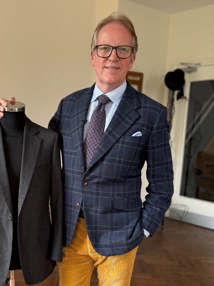 Thomas Mahon wearing a navy blue moonbeam check blazer, light blue dress shirt, patterned tie, and mustard yellow trousers stands indoors next to a black suit jacket displayed on a mannequin torso.